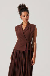 The woman is wearing a sleeveless brown blazer with a lapel collar and a matching belt tied at the waist. She pairs it with a long, pleated brown skirt.